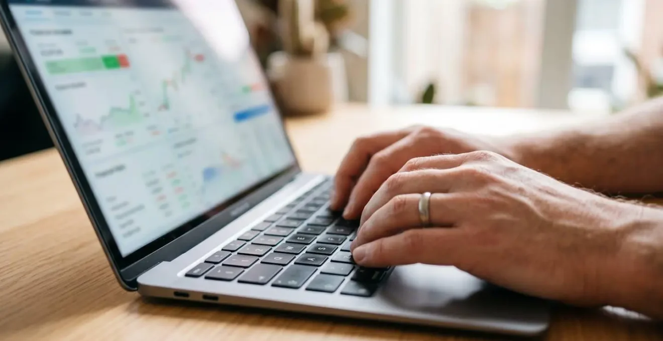 Gros plan sur des mains adultes tapant sur un clavier d'ordinateur portable moderne, interface de simulateur financier partiellement visible et floutée à l'écran